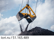 Heavy machinery is actively involved in unloading coal from a large bucket at an industrial location. The excavator crane, equipped with a dual clamshell bucket, hovers above a substantial pile of coal. Стоковое фото, фотограф Владимир Разгуляев / Фотобанк Лори