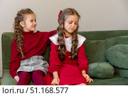 Two little girls sisters are sitting on a sofa near a Christmas tree. One girl is upset and cries, the other one hugs her and calms her down. Childhood and friendship relations concept. Стоковое фото, фотограф Ирина Михайличенко / Фотобанк Лори