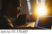 A man is seen working intently on a laptop in a dimly lit room, with the golden glow of the setting sun streaming through the window behind him. He wears glasses, emphasizing a focus on concentration and screen work. The atmosphere suggests late-night dedication or early-morning productivity, commonly associated with programming, development work, or other digital professions. The warm lighting and sharp silhouette create a dramatic contrast, reinforcing the mood of deep focus and solitude. This image evokes themes of remote work, tech culture, and the commitment often seen in freelance or startup environments. Стоковое AI-изображение, AI-художник Ксения Чунаева / Фотобанк Лори