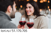 A couple sitting across from each other at a cozy, warmly lit restaurant. The woman is smiling, looking at her partner as they toast with glasses of red wine. Both are wearing winter sweaters, suggesting a romantic evening in a festive or cold-season setting. The soft bokeh lights in the background enhance the intimate atmosphere. Стоковое AI-изображение, AI-художник Ксения Чунаева / Фотобанк Лори