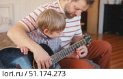 Handsome dad teaches his cute son to play the guitar sitting on sofa in the living room at home. Happy Father's Day. Father teaching his child to play guitar. Kid Learning music skill. Art education. Стоковое видео, видеограф Марина Демидюк / Фотобанк Лори