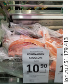Supermarket counter with chilled fish wrapped in transparent film. Large pieces of red fish - trout (kirjolohi) - stand out in the center of the frame, placed in a display case with a price tag. The sign says: "Kokonainen kirjolohi" (whole rainbow trout), country of origin - Finland (Suomi). The price is 10.90 euros per kilogram. The fish is presented both whole and cut into pieces, the fresh flesh of a bright orange color is visible. The atmosphere of the photo is typical for a grocery store, where the display cases are neatly decorated and emphasize the freshness of the product (2019 год). Редакционное фото, фотограф Анна Кривицкая / Фотобанк Лори