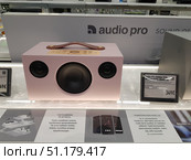 A modern portable speaker system Audio Pro Addon G10, presented in the window of an electronics store. The speaker has a stylish minimalist design with a soft pink body and a comfortable leather carrying handle. On the top panel there are control buttons and a rotary volume control in a gold design. The central part of the body contains a large speaker, on the sides there are two additional emitters. Next to the speaker you can see a price plate - the cost is 349 euros. At the bottom there is advertising information demonstrating the functions, including Bluetooth 4.0, the ability to synchronize with several speakers and control via a mobile application for iOS and Android. (2019 год). Редакционное фото, фотограф Анна Кривицкая / Фотобанк Лори