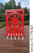 In the settlement of Nizino, which is located in the Lomonosovsky District of the Leningrad Region, not far from St. Petersburg. In the foreground is a modern decorative structure of bright red color, installed on a wooden platform. In the center of the composition is a circle with an eight-petal ornament, stylized as a solar sign, symbolizing the sun and harmony. At the bottom of the structure, the name of the settlement is written in white letters - Nizino. In the background, green trees, elements of a landscaped park and a pond are visible, which emphasizes the well-groomed and natural beauty of the rural settlement. Стоковое фото, фотограф Анна Кривицкая / Фотобанк Лори