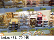 A refrigerated counter of a supermarket with a large assortment of confectionery products in transparent plastic packaging. On the shelves are various pastries, cakes, baskets with cream, profiteroles, pastries with berries, rolls and portioned desserts. Bright price tags indicating the cost are located under each type of product. In the center, pastries with white cream and sprinkles are noticeable, next to them are chocolate and biscuit desserts of the Mirel brands and other Russian manufacturers. The atmosphere of the photo conveys the usual selection of sweets in the store, designed for the convenience of customers. (2015 год). Редакционное фото, фотограф Анна Кривицкая / Фотобанк Лори