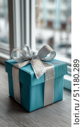 Gift Box Wrapped in Blue Paper With Silver Ribbon Sits on a Table by the Window. Стоковое AI-изображение, AI-художник Юлия Гапеенко / Фотобанк Лори