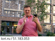 Dissatisfied millennial man in a pink T-shirt, chatting on the phone, looking at a gadget with an offended and disgusted expression, receiving a bad message. Стоковое фото, фотограф Данила Большаков / Фотобанк Лори