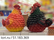 Two decorative rooster figurines, made in bright saturated colors. One of them is painted in red-orange-yellow shades, creating the effect of lush plumage with shimmering fiery tones. The second rooster has a black-green-red coloring with deep contrasting colors, giving it a more strict and elegant look. The figurines have an expressive relief in the form of scales-feathers, which adds to their decorativeness. Both items are placed on white boxes in the store, the background is blurred, creating an emphasis on the details of the toys.Such images can be used to illustrate holiday products, rooster symbols in decor, souvenirs and thematic compositions. Стоковое фото, фотограф Анна Кривицкая / Фотобанк Лори