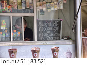 The window of a street kiosk or small shop. In the foreground hangs a chalk board with the inscription: "Currency is not sold, but accepted as a gift." Above the window are various banknotes from different countries of the world, used as a decorative element. Inside the kiosk, behind the glass, you can see bottles of water and fizzy drinks with price tags, as well as chocolate bars and juices. Behind the counter is a seller, intently looking at his phone. Below is part of an advertising banner with an image of coffee and ice cream. The atmosphere of the photo combines humor, everyday life and street trading. (2015 год). Редакционное фото, фотограф Анна Кривицкая / Фотобанк Лори