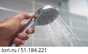 Close-up of hand holding chrome shower head with water flowing, in a modern tiled bathroom setting, showing daily hygiene and water use concept. Стоковое AI-изображение, AI-художник Катерина Бондарец / Фотобанк Лори