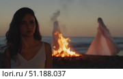 A young woman stands near a fire on the beach, while a ghostly figure appears in the background as the sun sets over the ocean. Стоковое AI-видео, аниматор Данила Большаков / Фотобанк Лори