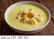 Cauliflower creamy soup with baked cauliflower florets on the top in a white bowl on a dark wood tabletop. Restaurant style dish. Hot, delicious plate of cream-of-cauliflower soup with melted cheese. Стоковое AI-изображение, AI-художник Марина Демидюк / Фотобанк Лори