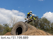 Magnitogorsk, Russian Federation - August 23, 2025: Motocross rider performs daring jump over large tire on dirt mound during enduro competition, showcasing skill and adrenaline in action-packed scene. Редакционное фото, фотограф Ирина Ручушкина / Фотобанк Лори