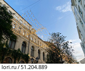 Moscow, Russia, July 22, 2025: view of Kuznetsky Most Street. Festive decoration lamps hang overhead historical street. Charming architecture features intricate details with greenery climbing. Редакционное фото, фотограф Olga Monako / Фотобанк Лори