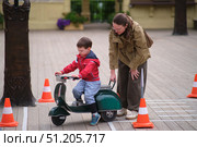 Woman helping a boy to ride a toy scooter. Kid learns to balance a scooter on a wooden path. Childhood education and parenting. A child aged 3 years. Стоковое фото, фотограф Андрей Журавлев / Фотобанк Лори