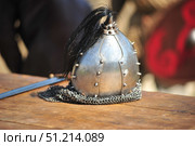 An antique metal helmet with spikes and a ponytail on top. The helmet is hemispherical in shape with segments connected with rivets and decorated with small metal spikes on the surface. A piece of chainmail is attached to the bottom to protect the neck. The helmet lies on a wooden table, with part of a sword blade visible nearby. In the background, details of the reconstruction are blurred - a shield with a red ornament and figures in historical costumes. The atmosphere of the photo conveys the spirit of the Middle Ages, military history and reenactment festivals. Стоковое фото, фотограф Анна Кривицкая / Фотобанк Лори