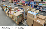 The process of displaying goods in a supermarket. In the foreground are wooden pallets with cardboard boxes, some of which are open. The boxes contain packages of milk, yogurt and other dairy products. A trolley for moving boxes is visible. In the background is a retail shelving unit with a large assortment of dairy products: milk, cream, kefir, yogurts in cardboard packages of different brands. The atmosphere of the photo conveys the work process of the store, the organization of supplies and logistics. Редакционное фото, фотограф Анна Кривицкая / Фотобанк Лори