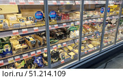 A refrigerated display case in a supermarket with a large assortment of cheeses. The shelves display various types: hard cheeses, parmesan, slices, cheeses in bars, pieces and grated varieties. You can see the packaging of both domestic and imported brands, neatly laid out in boxes and individual packages. Some of the products are in transparent film, others are in branded packages with bright labels. The display case is closed with glass doors with price tags and advertising stickers. The atmosphere of the photo conveys the abundance of choice in a modern supermarket. Редакционное фото, фотограф Анна Кривицкая / Фотобанк Лори