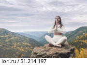 White woman practices yoga in lotus position while meditating on rocky cliff surrounded by autumn mountains. Yoga practice and outdoor fitness. Стоковое фото, фотограф Владимир Разгуляев / Фотобанк Лори