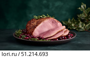 A beautifully roasted ham is elegantly sliced and presented on a dark ceramic plate, garnished with fresh rosemary sprigs and surrounded by plump red cranberries. The ham's succulent pink interior contrasts with its perfectly browned, seasoned crust, highlighting expert preparation. Set against a dark green backdrop with soft holiday greenery, the composition evokes a festive, cozy, and traditional dining atmosphere. The overall mood is warm and celebratory, making this image ideal for seasonal food promotions, holiday recipe features, or culinary editorials focused on comfort food, festive cooking, and classic main courses. Стоковое AI-изображение, AI-художник Ксения Чунаева / Фотобанк Лори