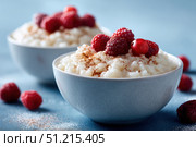 Milky Rice pudding with superberries and red raspberry sauce, served in a white bowl on blue tablecloth background. Rice porridge topped with some raspberries and cinnamon sprinkled. Old English style. Стоковое AI-изображение, AI-художник Марина Демидюк / Фотобанк Лори