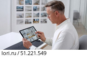 Mature architect reviewing various house project drafts on a digital tablet, working in a modern office with design samples displayed on the wall. Стоковое AI-изображение, AI-художник Юрий Поздников / Фотобанк Лори