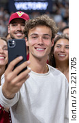 Young man taking selfie with friends at stadium, celebrating team victory, surrounded by cheering crowd, expressing raw excitement and camaraderie. Стоковое AI-изображение, AI-художник Юрий Поздников / Фотобанк Лори