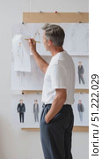 Professional fashion designer with gray hair drawing tailored menswear sketches, carefully refining suit designs mounted on studio workspace board. Стоковое AI-видео, аниматор Юрий Поздников / Фотобанк Лори
