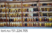 A honey shop where jars with different varieties of this natural product are neatly displayed on wooden shelves. Glass jars of different sizes — from small 0.25 l to large three-liter ones — are filled with honey of all possible shades: from almost white to dark brown. The varieties are visible: buckwheat, linden, sweet clover, hawthorn and others. There are price tags under each row. The interior is dominated by wooden shelves, creating an atmosphere of home comfort. On some shelves you can also see nuts in honey, herbal infusions and souvenirs, which gives the room the flavor of a farmer's shop. Стоковое фото, фотограф Анна Кривицкая / Фотобанк Лори