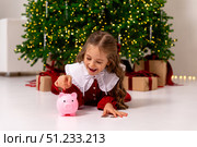 Banking, finances, investment and happy people concept. Cute happy little girl lies a floor and puts money coins in pink piggy bank on Christmas. Pocket money, family budget and savings concept. Стоковое фото, фотограф Ирина Михайличенко / Фотобанк Лори