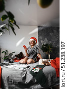 Woman in Santa hat enjoys Christmas at home, using smartphone for communication and social media. With help of modern technology, celebrates Christmas by sharing holiday moments and staying connected. Стоковое фото, фотограф Енваев Андрей / Фотобанк Лори