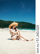 Peaceful summer scene: young woman in straw hat sits barefoot on sand, looking thoughtfully toward sea moment of solitude and connection with nature. Стоковое фото, фотограф Енваев Андрей / Фотобанк Лори