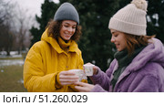 Two friends share warm drinks while wearing cozy winter outfits in a snowy park during a chilly afternoon. Стоковое AI-изображение, AI-художник Сергей Толмачёв / Фотобанк Лори