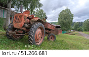 Old red rusty russian tractor t 16 parked beside gravel road in rural area surrounded by trees and old buildings during a cloudy afternoon. Стоковое фото, фотограф Владимир Разгуляев / Фотобанк Лори