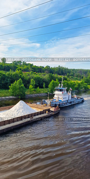 A barge with a cargo of white rubble is moving along the Volga River, which is being dragged by a small tugboat with a Russian flag against the background of a green forest. Стоковое фото, фотограф Анна Чижова / Фотобанк Лори