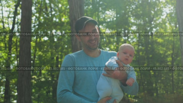 A bearded father in a blue jersey and black baseball cap holds a baby in a blue onesie against a forest background. A man smiles while playing with his son in the park holding him in his arms. Стоковое видео, видеограф Воловодов Владимир / Фотобанк Лори