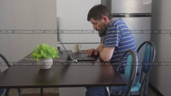 A bearded man is working on a laptop in the kitchen at a black table. Стоковое видео, видеограф Ярославский Максим / Фотобанк Лори