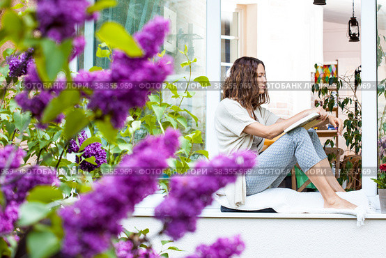 A beautiful middle-aged woman with a book in her hands sits on the windowsill. Стоковое фото, фотограф Astroid / Фотобанк Лори