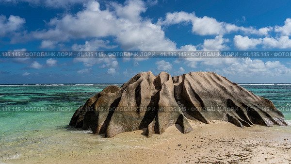 A bizarre granite boulder with smoothed folded slopes on the beach of a tropical island. The waves of the turquoise ocean spread over the wet sand. Clouds in the blue sky. Seychelles. La Digue. Стоковое фото, фотограф Вера Ивановна Кубарева / Фотобанк Лори