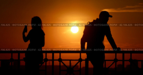 A blurred photograph of two people silhouettes a man and a woman at sunset in the city. A woman takes a selfie, a man in a cap looks away. Стоковое фото, фотограф Валерий Леонидович / Фотобанк Лори
