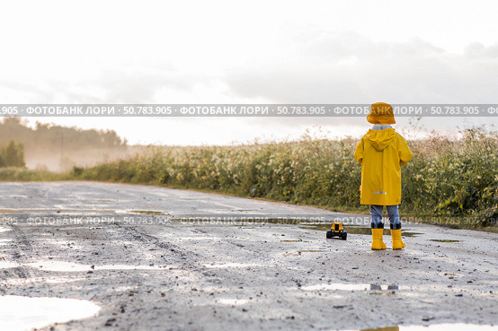 A boy in yellow rubber boots and a raincoat runs through puddles, having fun and playing with a remote-controlled car after it rains. Summer and autumn holidays. A happy childhood. Стоковое фото, фотограф Photojuli / Фотобанк Лори