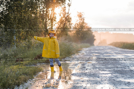 A boy in yellow rubber boots and a raincoat runs through puddles, having fun and playing after it rains. Summer and autumn holidays. A happy childhood. Стоковое фото, фотограф Photojuli / Фотобанк Лори