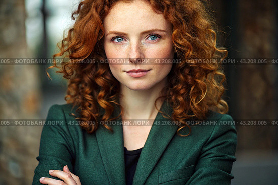 A businesswoman with curly red hair and freckles on her face. She is dressed in a casual dark green blazer and jacket. Arms crossed over his chest, looking straight at the camera. Стоковое AI-изображение, AI-художник Photojuli / Фотобанк Лори