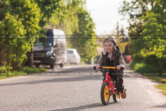 A cheerful little boy rides a bicycle outdoors. A happy child walks in the spring park. The baby is dressed in a fashionable vest, overalls. An aviator's hat is on his head. Стоковое фото, фотограф Photojuli / Фотобанк Лори