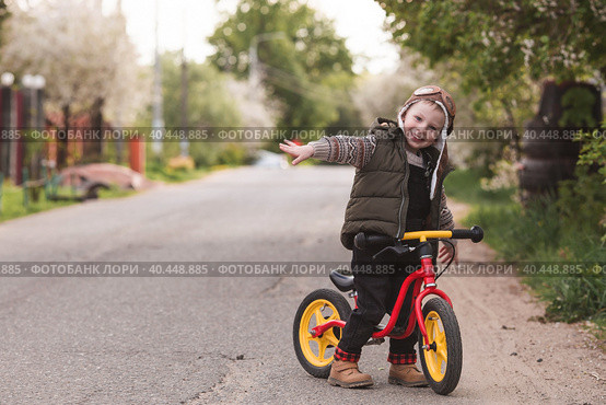 A cheerful little boy rides a bicycle outdoors. A happy child walks in the spring park. The baby is dressed in a fashionable vest, overalls. An aviator's hat is on his head. Стоковое фото, фотограф Photojuli / Фотобанк Лори