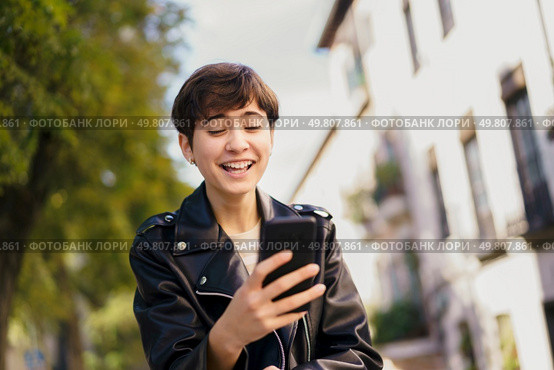 A cheerful young individual interacts with their smartphone against a colorful backdrop of modern architecture, embodying the essence of connectivity and communication in todays fastpaced world. A Young Individual Is Enjoying Their Mobile Phone While In An Urban Setting Today. Стоковое фото, агентство Ingram Publishing / Фотобанк Лори