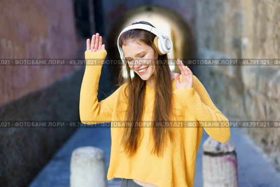 A cheerful young woman dressed in a bright yellow sweater and wearing headphones joyfully dances in a vibrant alley, fully embracing and celebrating the sheer joy and freedom that music brings. A Joyful Young Woman Is Enjoying Listening to Music While Outdoors and Loving Life. Стоковое фото, агентство Ingram Publishing / Фотобанк Лори