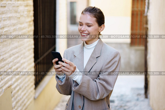 A cheerful young woman dressed in a chic outfit is engaging with her smartphone while walking through a charming alley. A cheerful young woman in a stylish outfit smiles while using her smartphone in a city alley. Стоковое фото, агентство Ingram Publishing / Фотобанк Лори