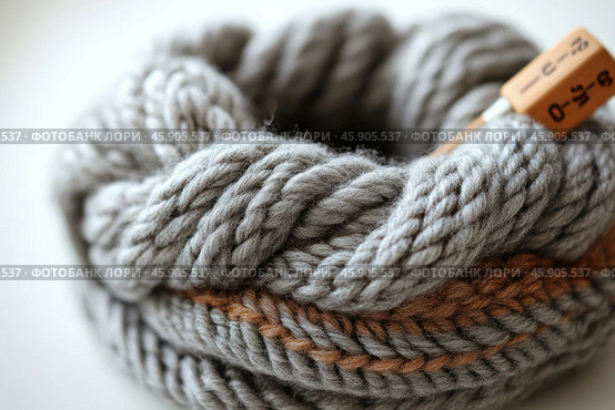 A close-up of a skein of thick gray yarn is shown. The knitting is made of soft and fluffy wool, with thick weaves visible, making it ideal for creating cozy items such as sweaters, scarves, or blankets. A contrasting brown stripe runs down the center of the skein, adding an accent. On the right is a wooden tag or label with numbers printed on it, which may indicate the length or composition of the yarn. The background is blurred, drawing attention exclusively to the texture and details of the knitting. Стоковое AI-изображение, AI-художник Анна Кривицкая / Фотобанк Лори