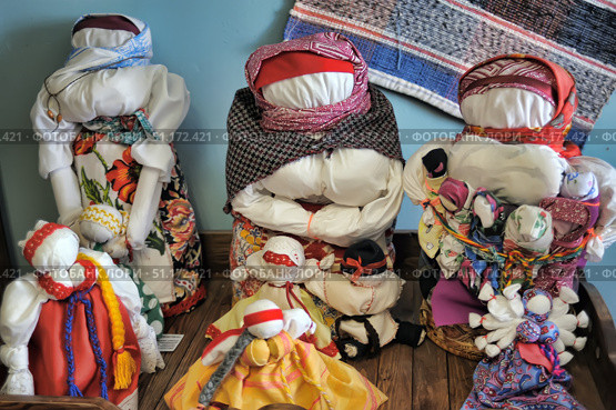 A collection of traditional Russian folk dolls-amulets, handmade from fabric. The dolls have no face, which corresponds to ancient traditions, symbolizing protection and protective power. Each figure is dressed in bright sarafans, shawls and aprons with ornaments and floral patterns. The photo shows dolls of different sizes - from large mother figures to small children's and family ones. On the back wall there is a woven rug, emphasizing the atmosphere of village life and ethnographic collection. The atmosphere of the photo conveys cultural heritage, folk traditions and the beauty of handicraft. Стоковое фото, фотограф Анна Кривицкая / Фотобанк Лори